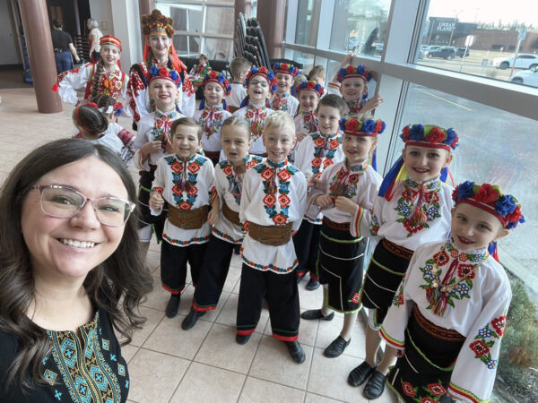 A group of children in colorful, traditional Ukrainian costumes smile and pose indoors. A woman in similar attire is taking a selfie with them.