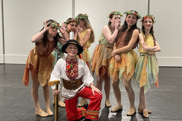 A group of children dressed in colorful Ukrainian costumes pose happily in a dance studio. A boy kneels in front, holding a wooden stick, surrounded by girls wearing floral crowns and vibrant dresses.