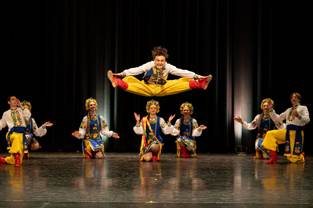 Boyan Dance Recital 2025 A Boyan dancer in vibrant traditional Ukrainian attire leaps energetically, legs spread wide, above seven kneeling performers on stage, radiating joy and energy.