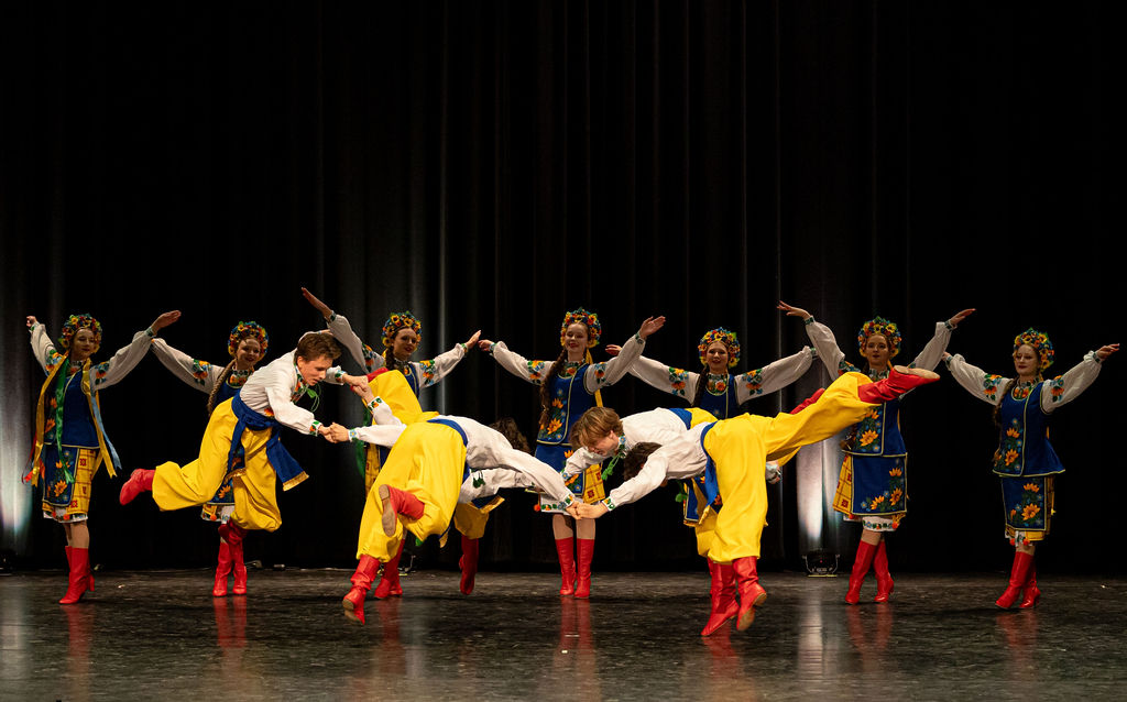 Boyan Dance Recital 2025 A group of dancers in traditional, colorful attire perform a Ukrainian folk dance on stage. They wear embroidered vests, yellow pants, and red boots, exuding joy and energy.