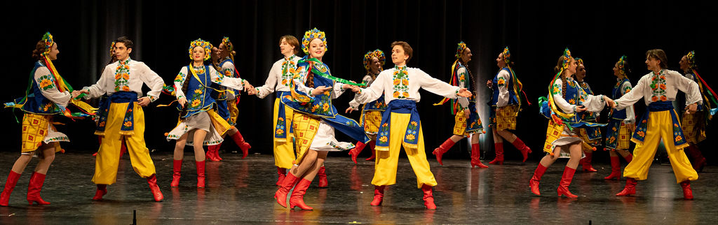 Boyan Ukrainian dancers in vibrant traditional costumes perform on stage with colorful embroidery, yellow pants, blue sashes, and red boots.