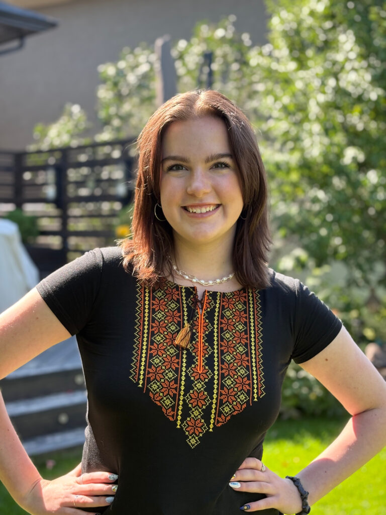 Anna Donahue Young woman smiling brightly in a sunlit garden, wearing a black shirt with colorful, intricate embroidery. Her hands are on her hips, exuding confidence.
