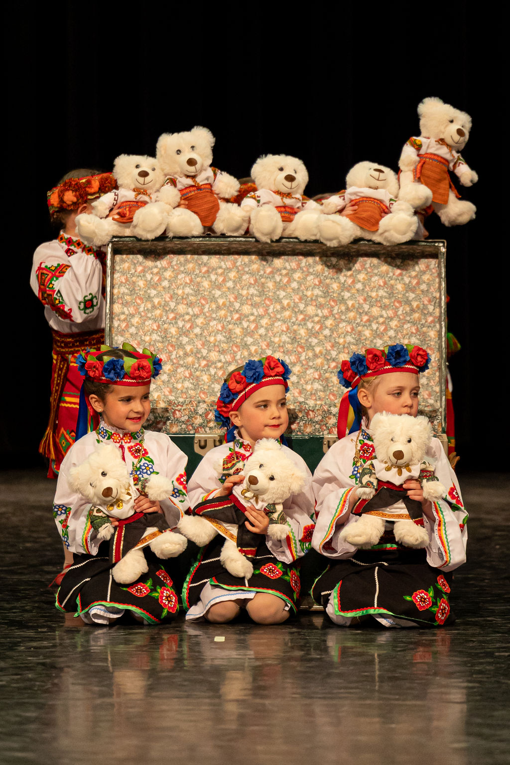 Three young children in colorful traditional Ukrainian outfits sit on stage holding teddy bears. A row of similar bears is displayed on a patterned chest behind them.