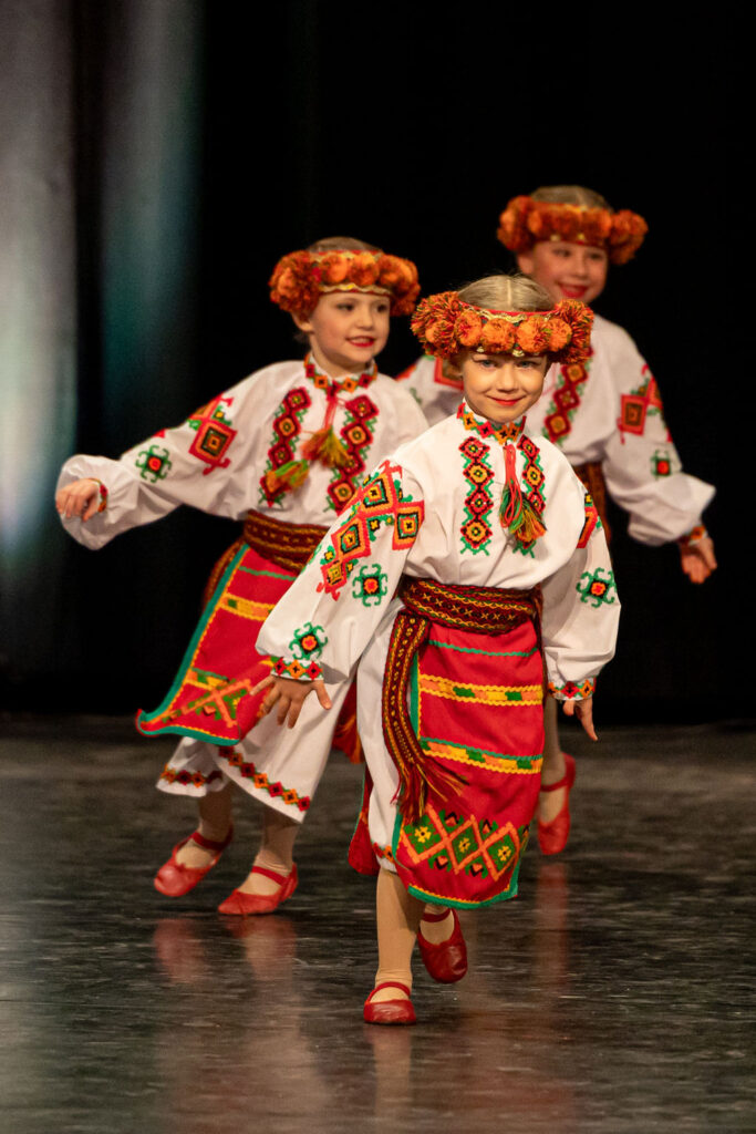 Boyan Dance Recital 2025 Children in traditional Ukrainian folk costumes dance joyfully on stage. They wear embroidered shirts, vibrant red skirts, and floral headpieces.
