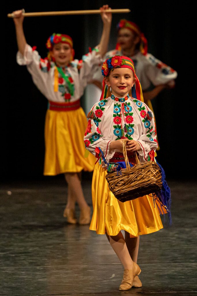 Boyan Dance Recital 2025 Young Ukrainian dancers in vibrant traditional attire perform on stage. The lead holds a woven basket, while others pose with wooden sticks.