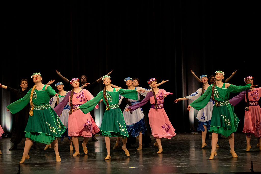 Boyan Dance Recital 2025 A group of Boyan dancers in vibrant traditional Ukrainian costumes, featuring green, pink, and blue, perform joyfully on stage. Their arms are outstretched in harmony.