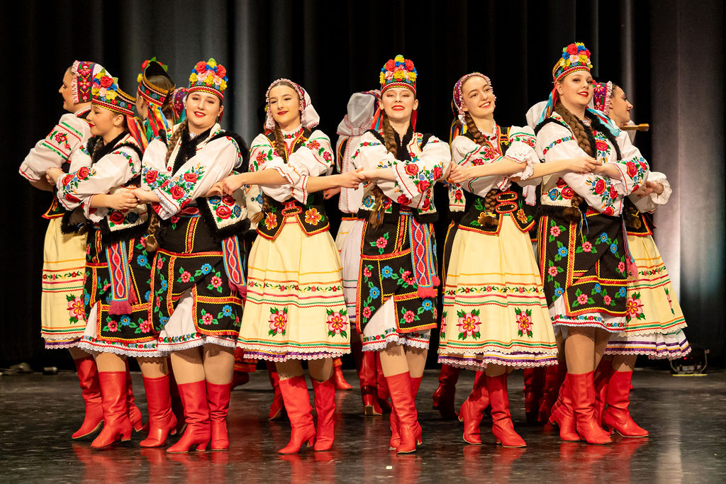 A group of Boyan dancers performs in vibrant traditional Ukrainian costumes, featuring colorful embroidery and red boots, against a dark stage backdrop. The scene conveys joy and cultural celebration.