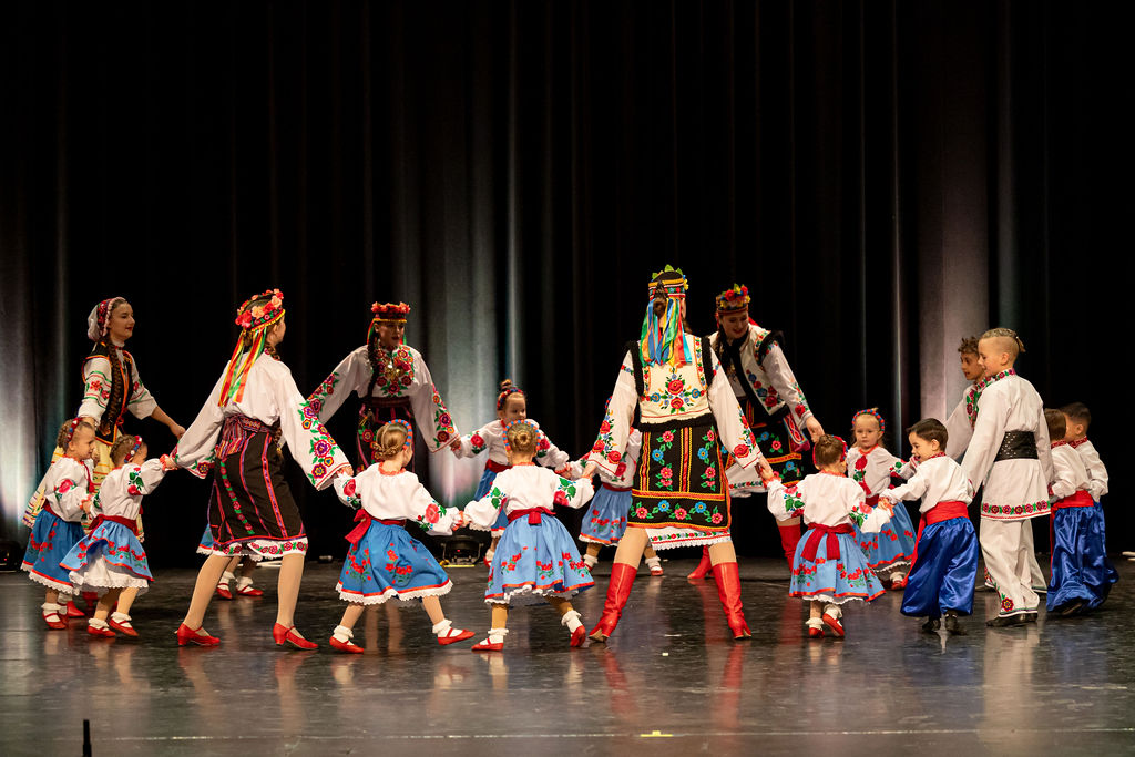 Children and adults in colorful Ukrainian dancing costumes dance in a circle on stage. The scene is lively and joyful, emphasizing cultural celebration.