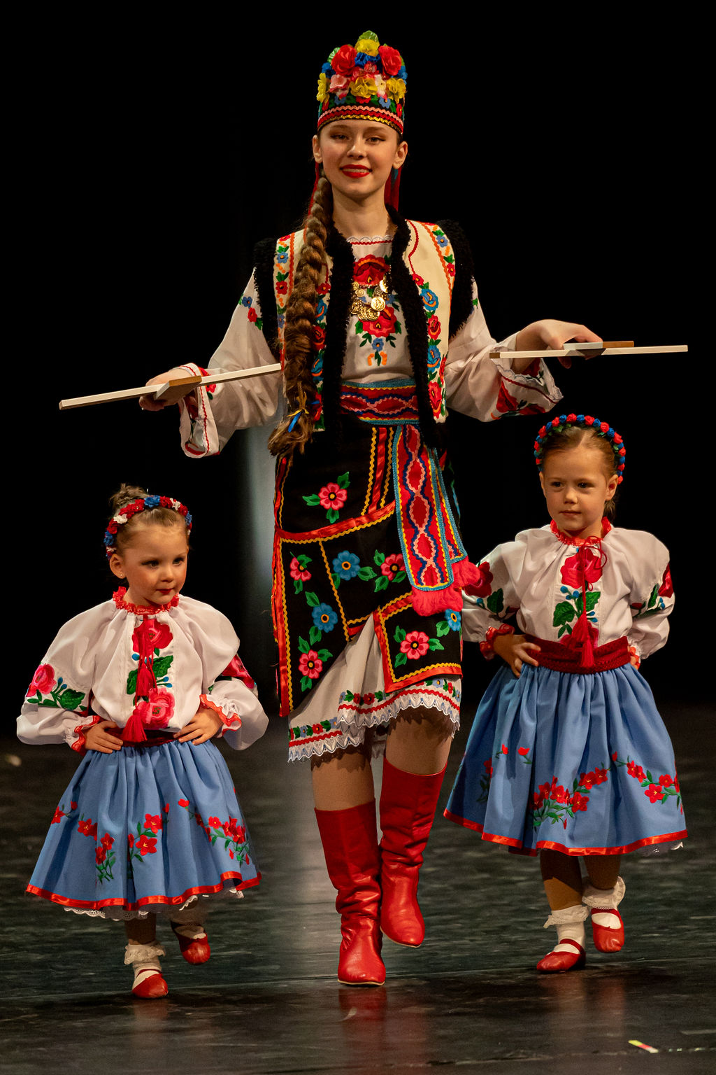 A woman and two young girls in vibrant traditional Ukrainian dancing outfits perform a cultural dance on stage. They wear colorful floral dresses and red boots.