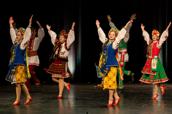 Boyan dancers in colorful traditional Ukrainian clothing perform on stage, hands raised. Bright floral patterns and vibrant colors create a lively, joyous atmosphere.