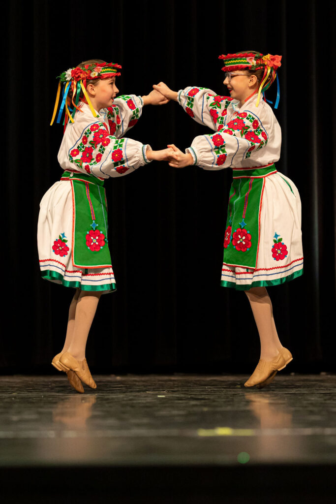 Boyan Dance Recital 2025 Two young dancers in traditional Ukrainian attire perform on stage. They wear embroidered outfits with colorful ribbons, holding hands joyfully.