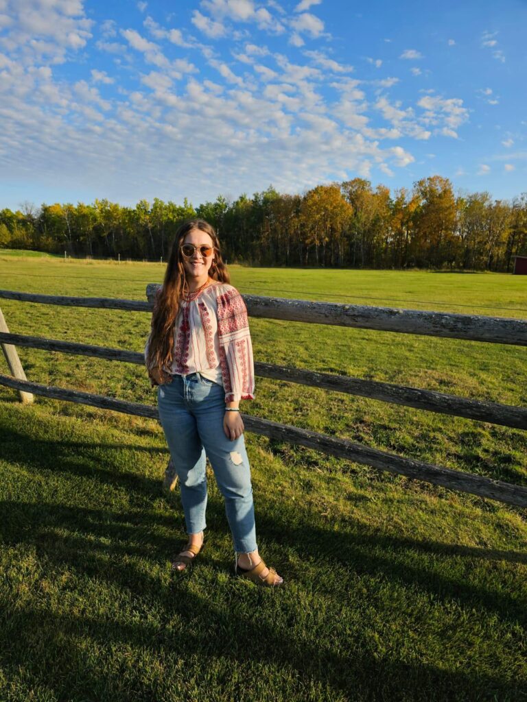 Natalya Shevchuk A woman with long hair and sunglasses stands by a wooden fence in a sunny field, wearing a patterned top and jeans. Autumn trees and a blue sky are in the background.